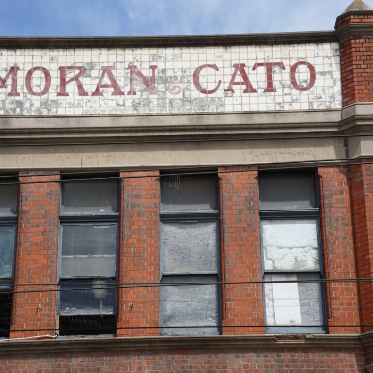 Faded Moran & Cato lettering on a tiled parapet above a red brick corner building, Melbourne inner north, hidden__north documentary photography