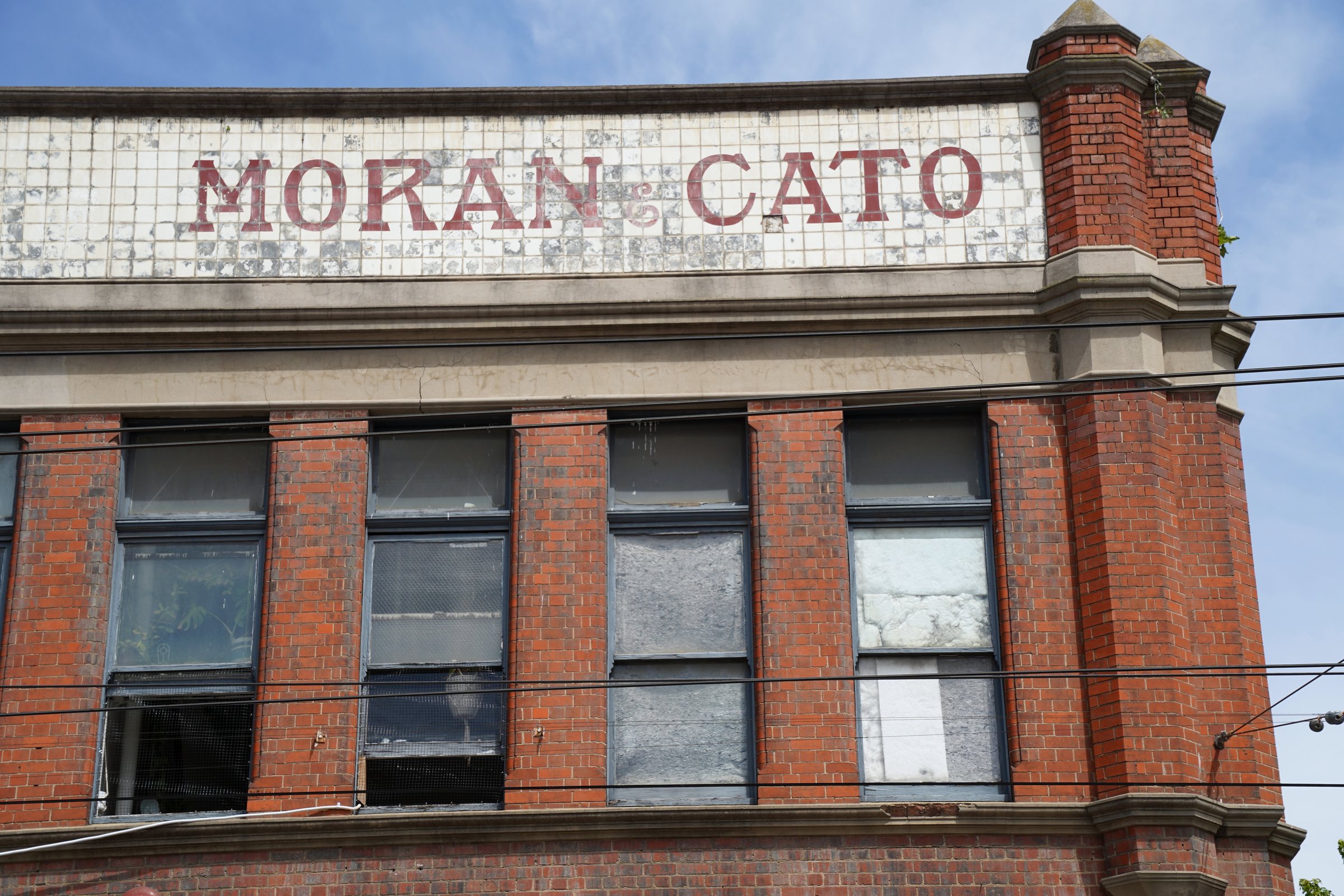Faded Moran & Cato lettering on a tiled parapet above a red brick corner building, Melbourne inner north, hidden__north documentary photography