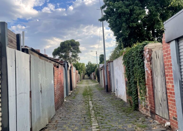 Cobbled laneway lined with garage doors and fences under a blue sky, Melbourne inner north, hidden__north documentary photography