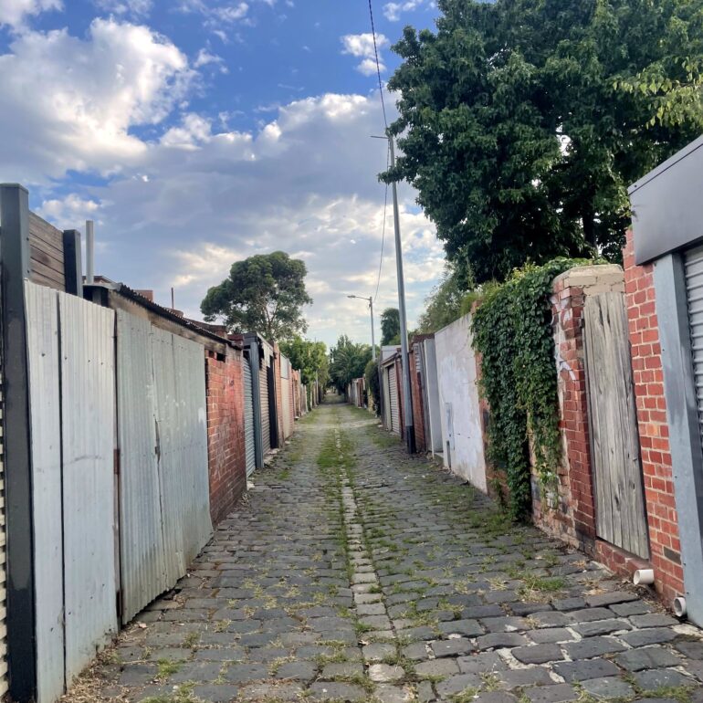 Cobbled laneway lined with garage doors and fences under a blue sky, Melbourne inner north, hidden__north documentary photography