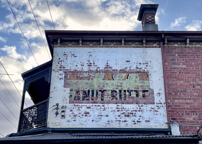 Faded painted ghost sign on a white rendered wall above a verandah, red brick corner building, Melbourne inner north, hidden__north documentary photography