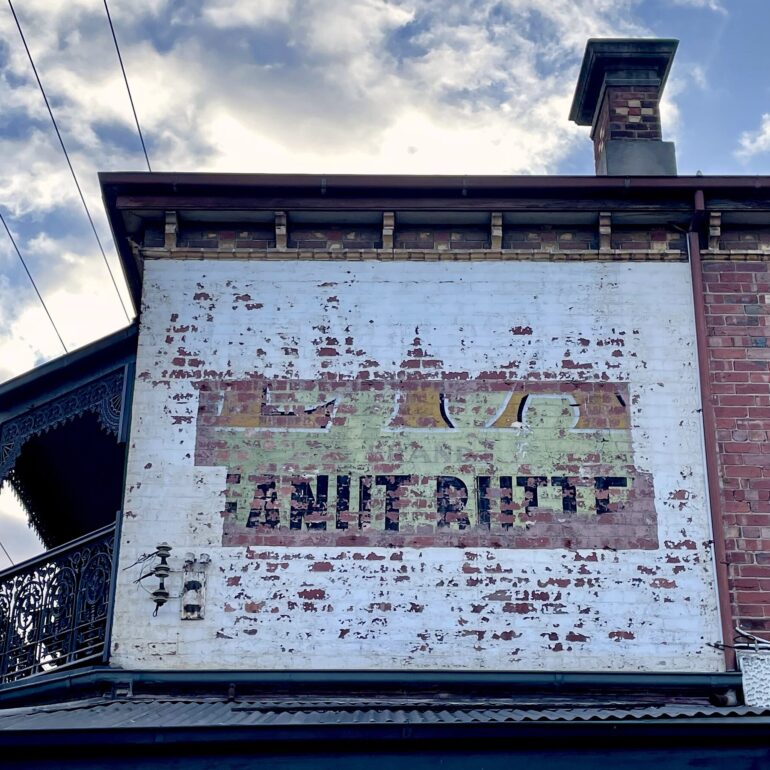 Faded painted ghost sign on a white rendered wall above a verandah, red brick corner building, Melbourne inner north, hidden__north documentary photography
