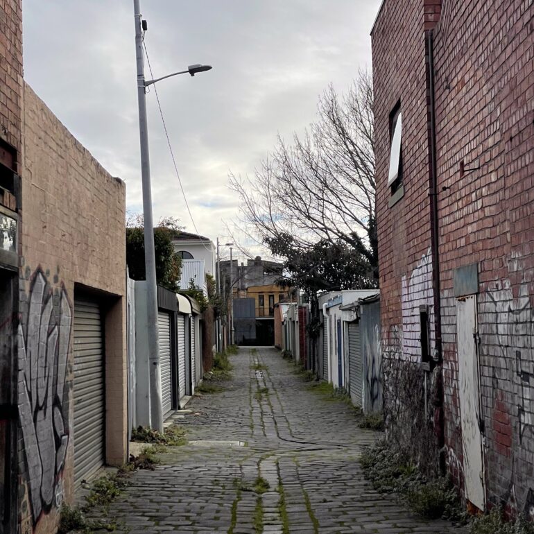 Narrow cobbled laneway lined with garage doors under an overcast sky, Melbourne inner north, hidden__north documentary photography